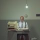 A man sits at a sad desk in a darkened basement with an old computer